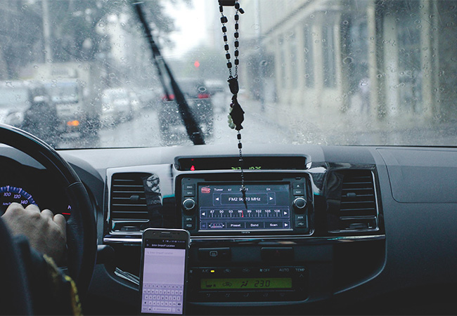 Blick auf ein Cockpit im Auto; auf dem Touchscreen ist die Radiofrequenz zu sehen und ein Handy hängt in einer entsprechenden Halterung neben dem Lenkrad auf Höhe der Hand des Fahrers. Bild: Pexels.com/Mary Whitney